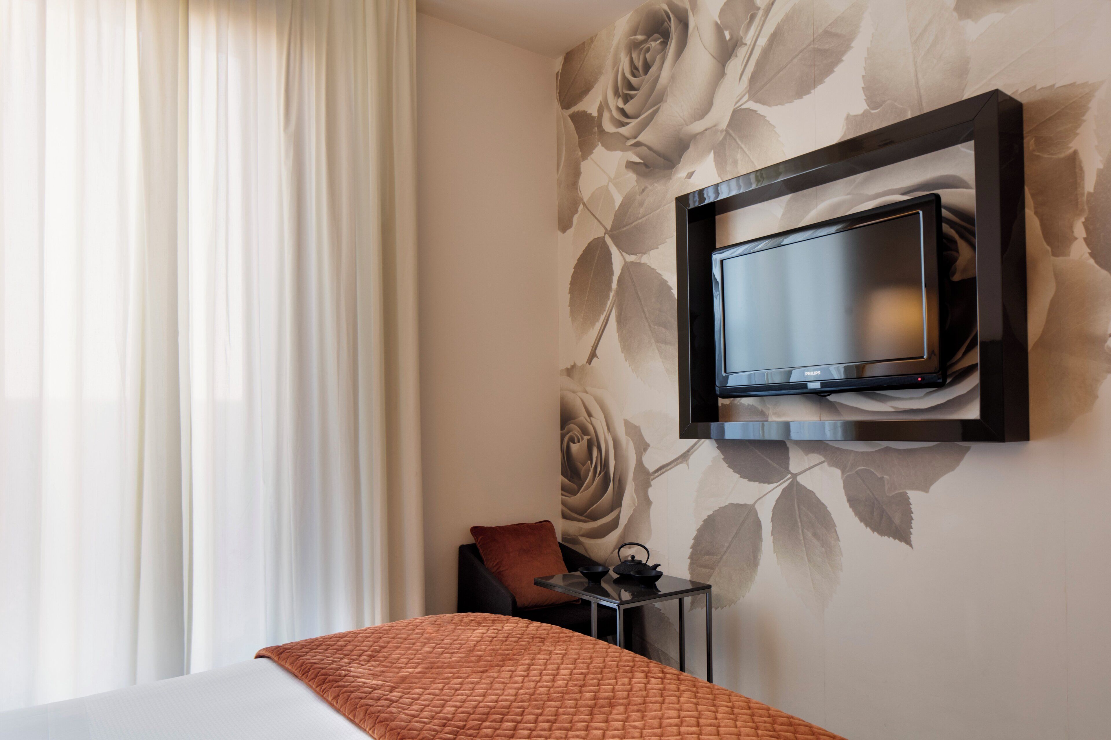 A hotel room with a bed, a TV mounted on the wall, a small table with a kettle, and a chair.