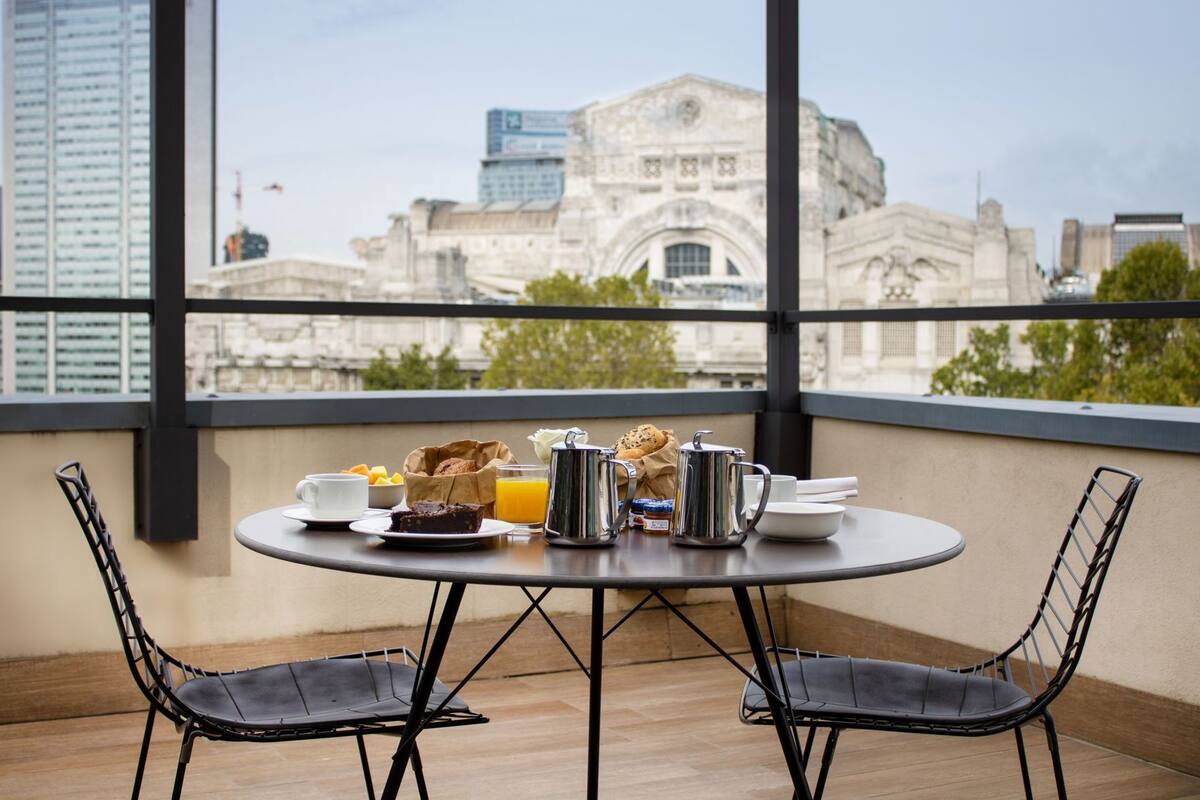 Une table ronde avec des aliments pour le petit-déjeuner, deux chaises et une vue sur des bâtiments d’architecture historique.