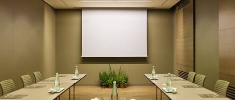 A conference room with a large screen, long tables, chairs, water bottles, and small plants.