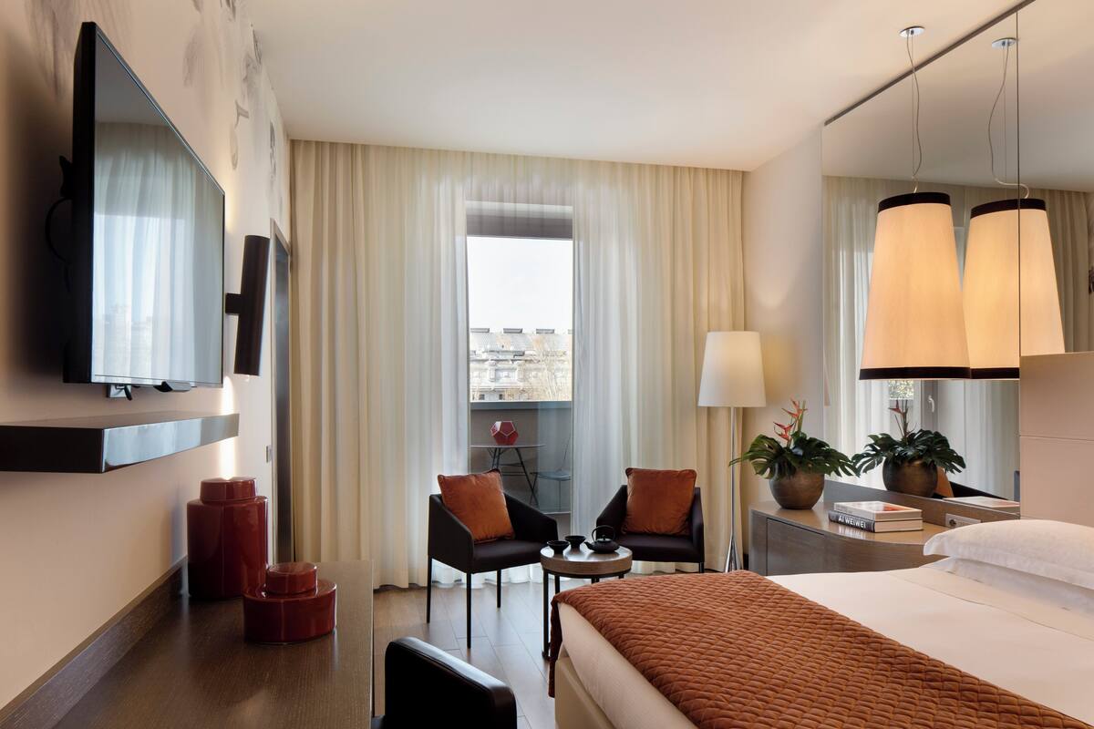Une chambre d’hôtel comprenant un lit, deux fauteuils, une petite table, un téléviseur, une fenêtre avec des rideaux et une plante en pot.