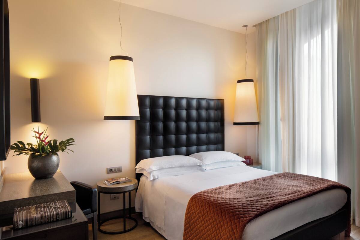 Une chambre d’hôtel avec un grand lit, un bureau avec une lampe, une petite table et une plante en pot.