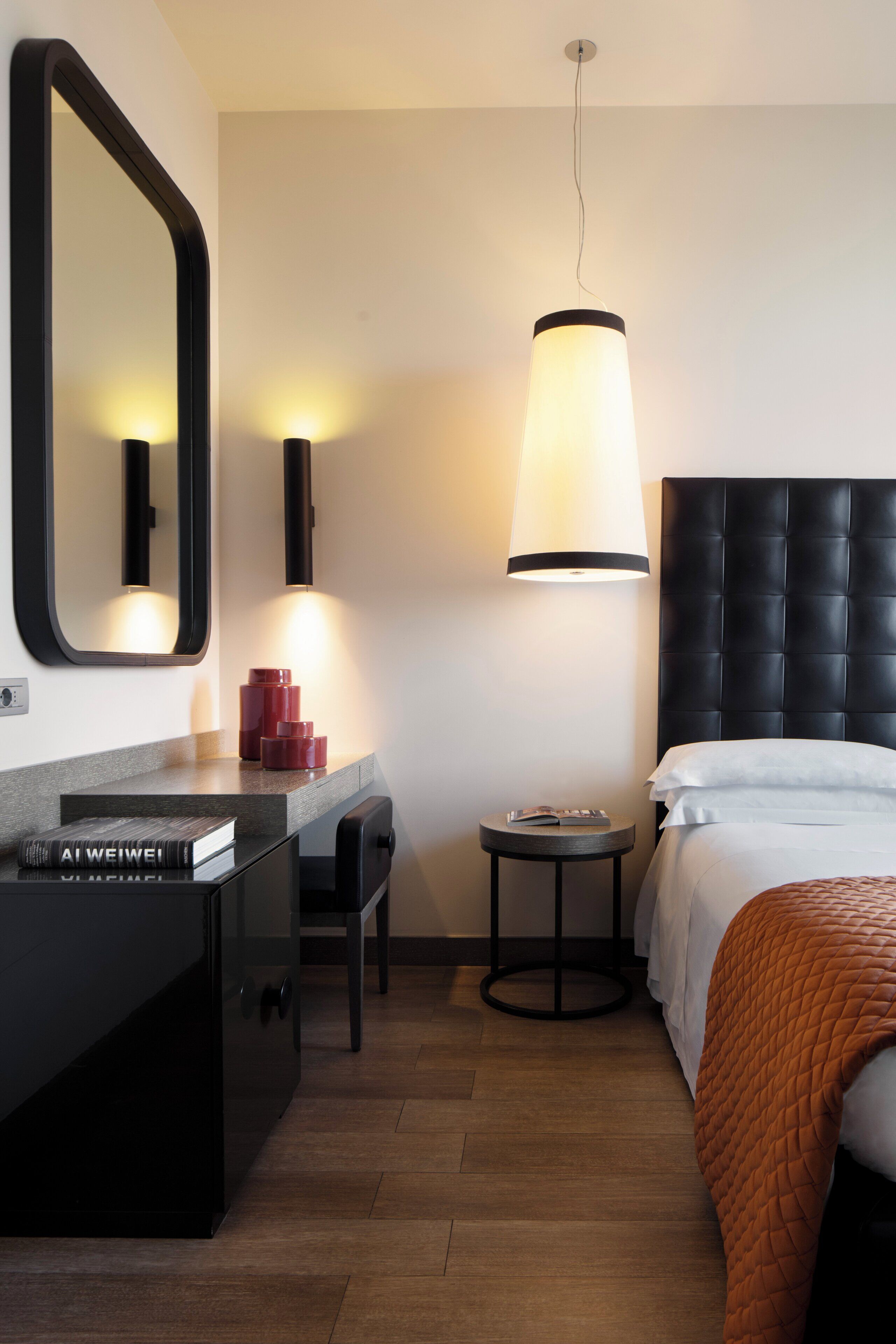 A hotel room with a bed, a bedside table with a lamp, a mirror, a small round table, and a wardrobe.