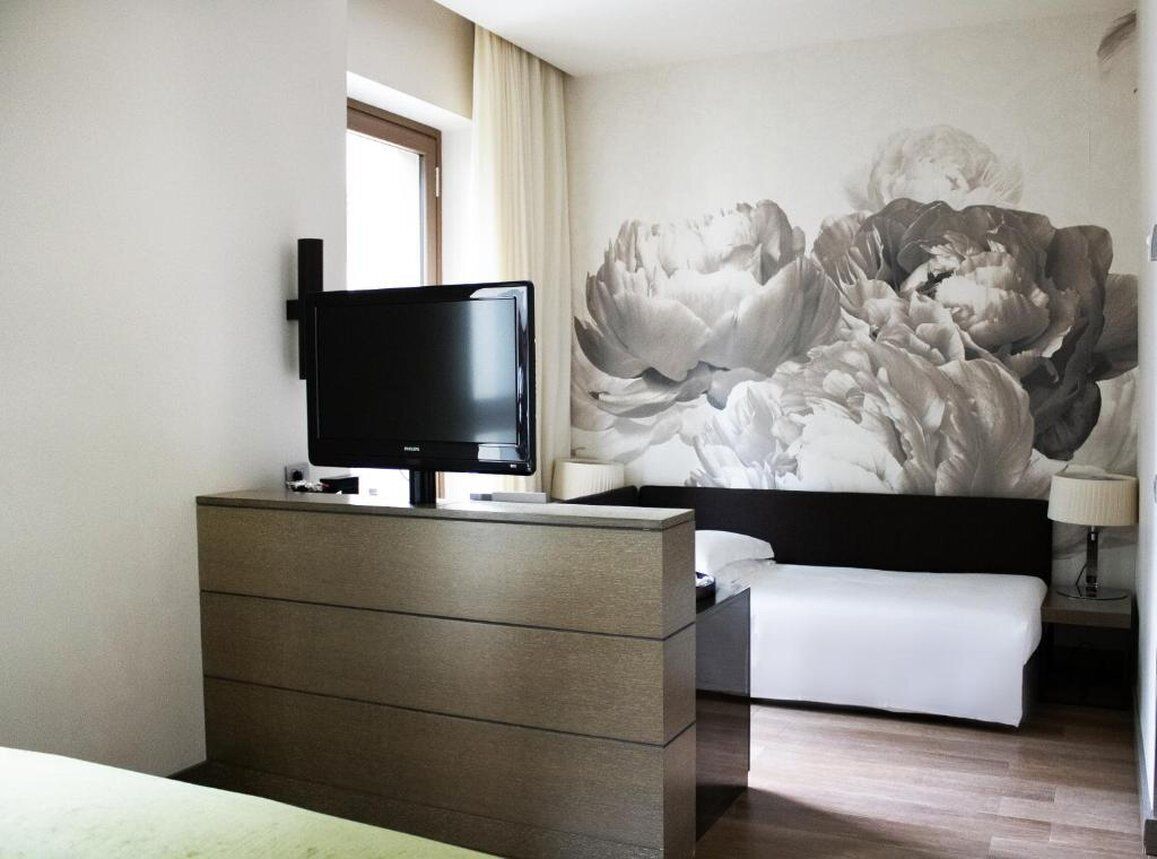Een hotelkamer met een bed, een televisie op een dressoir, een grote bloemenmuur en een raam met gordijnen.
