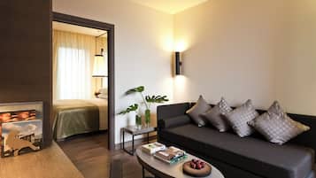 Une chambre d’hôtel moderne avec un canapé gris foncé, une petite table d’appoint ornée d’une plante, un tableau encadré et une table basse sur laquelle sont posés des livres et des fruits.