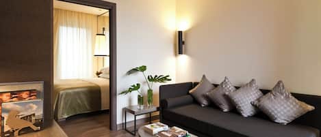 Habitación de hotel moderna con un sofá gris oscuro, una mesita auxiliar con una planta, un cuadro enmarcado y una mesa de centro con libros y fruta.