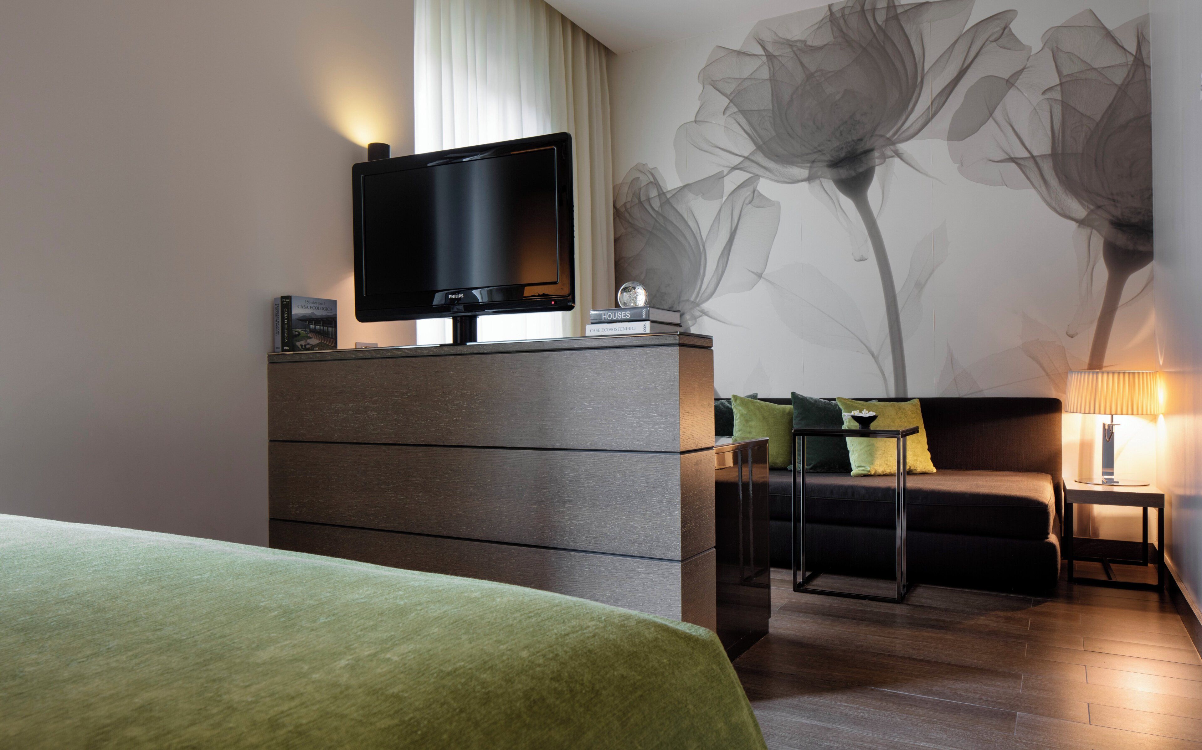 Une chambre d’hôtel moderne avec un téléviseur à écran plat posé sur une commode en bois, une grande fresque florale sur le mur, une couvre-lit verte et une table de chevet avec une lampe.