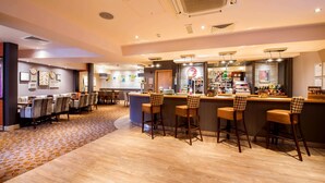A bar area with wooden bar stools, a counter with various items, and a seating area with tables and chairs. - Premier Inn London Victoria (London)