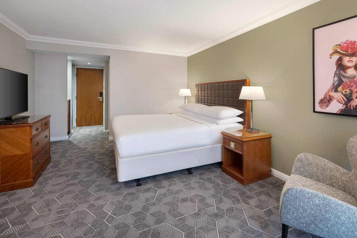 A hotel room with a bed, bedside tables, a TV, a chair, and a painting on the wall.