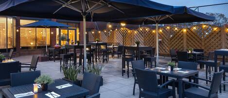 An outdoor patio area with tables and chairs, covered by umbrellas, and a wooden fence in the background.