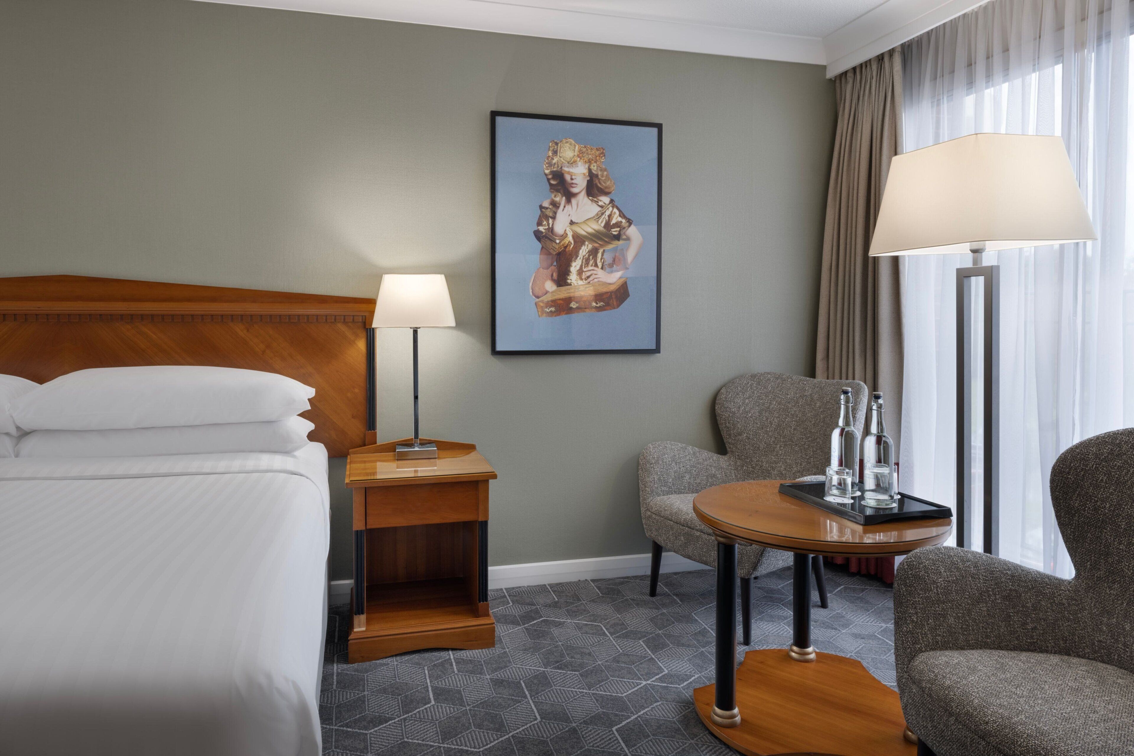 Una habitación de hotel con cama, mesita de noche, lámpara, silla y una mesa con botellas.
