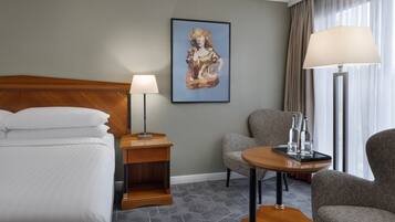 A hotel room with a bed, a nightstand, a lamp, a chair, and a table with bottles.