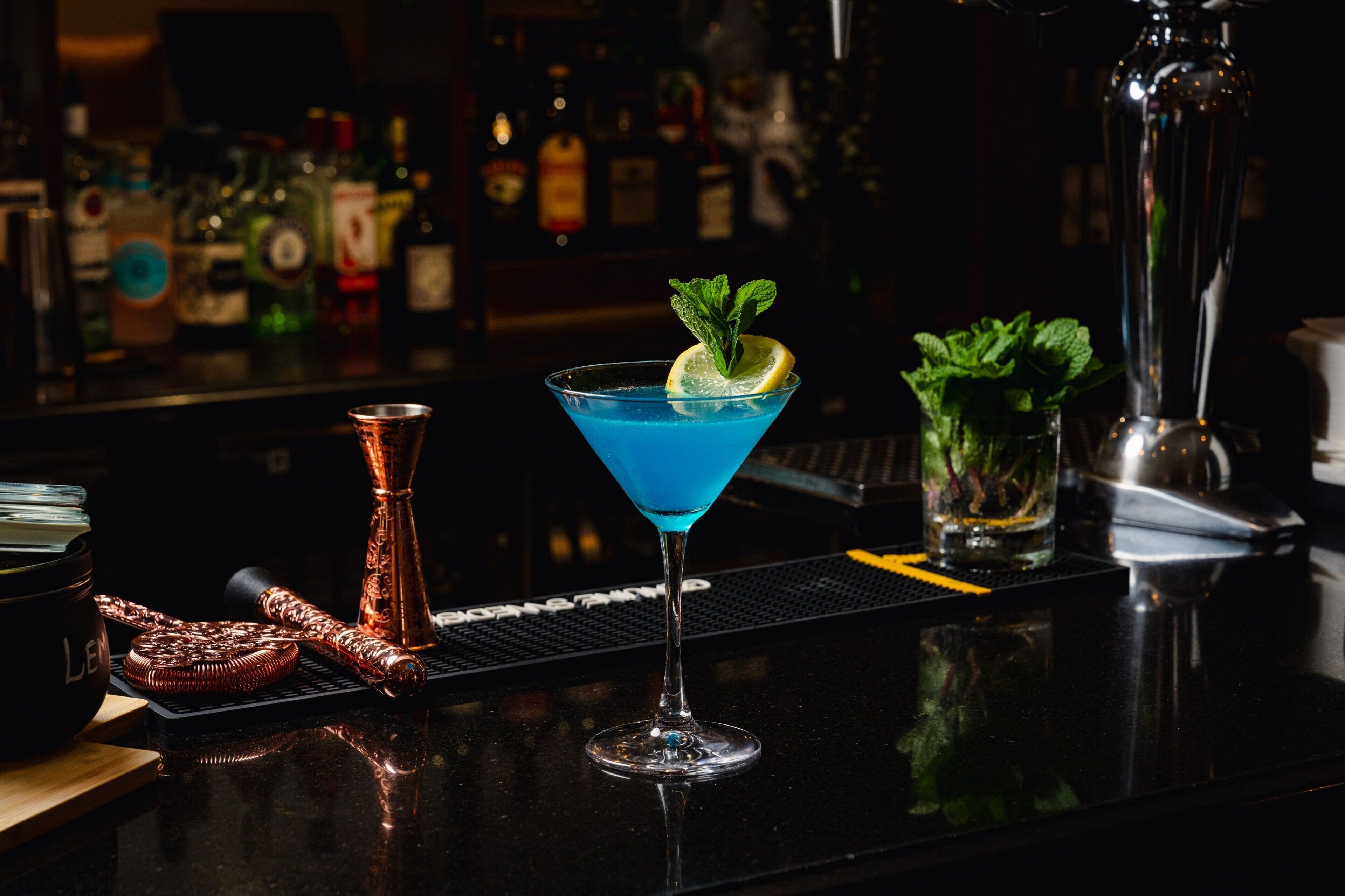 Un bar con un cóctel azul, un agitador de cobre y una ramita de menta.