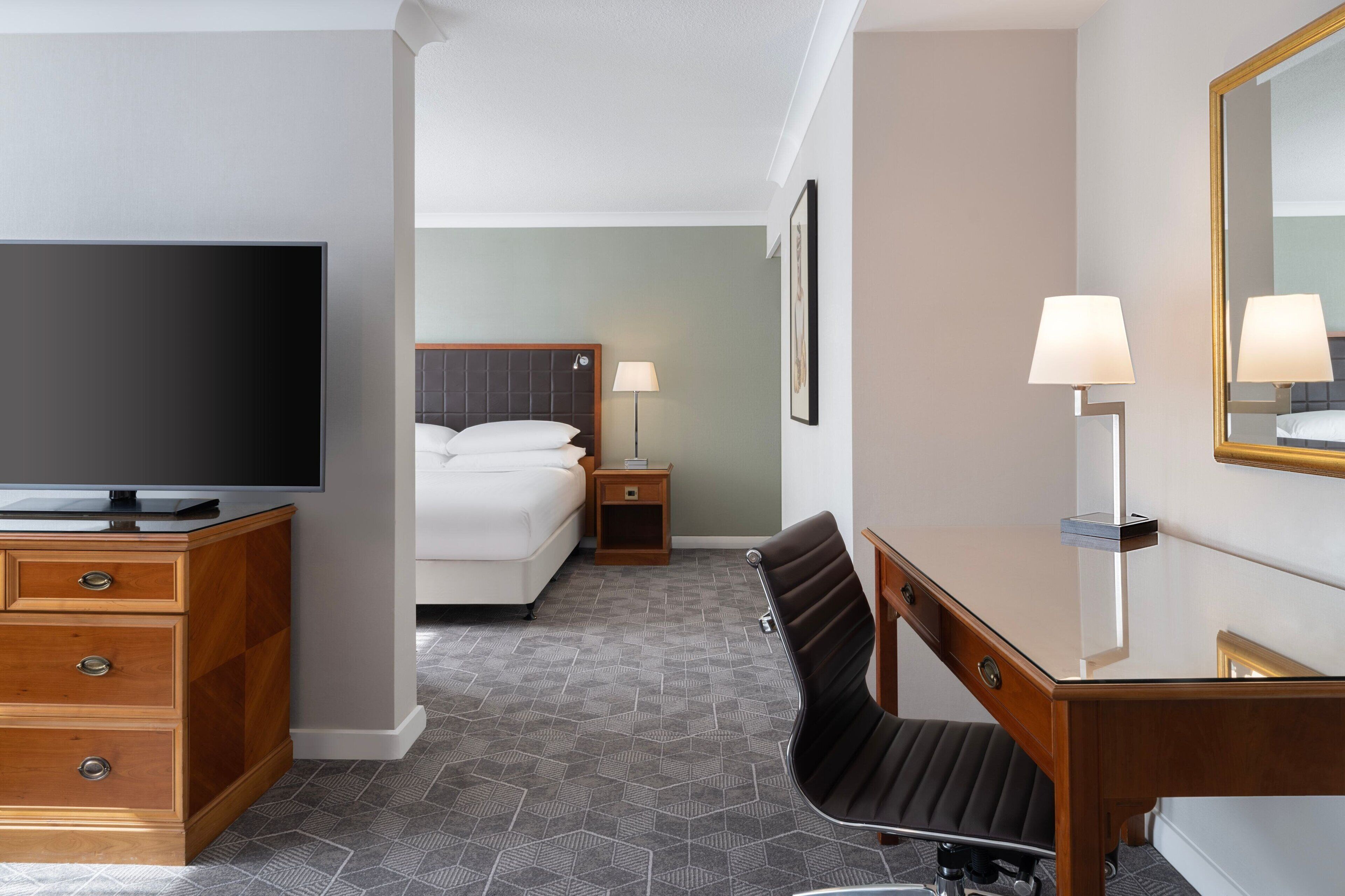 A hotel room with a bed, a desk with a chair, a television, a lamp, and a mirror.