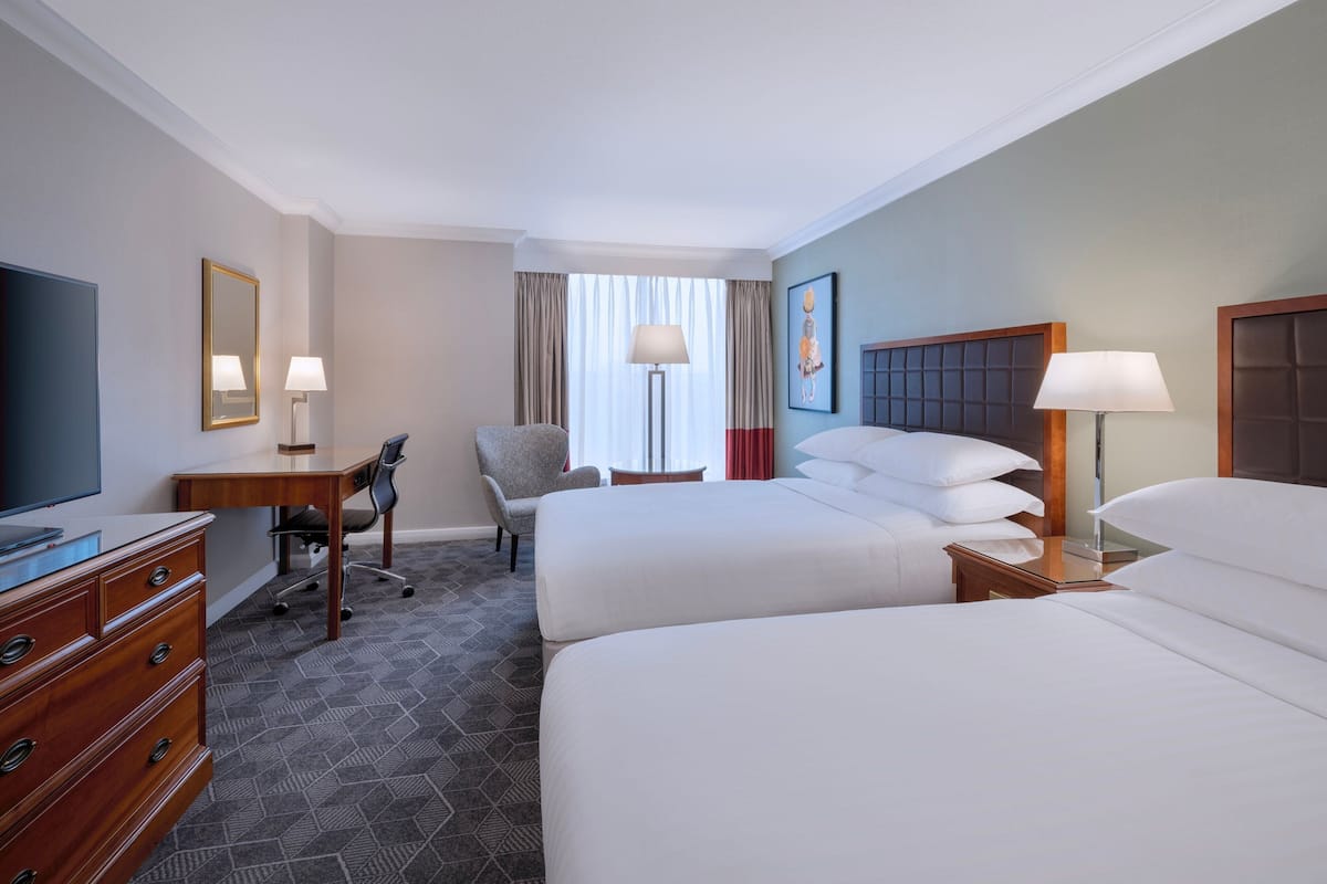A hotel room with two beds, a desk, a chair, a TV, and a lamp.