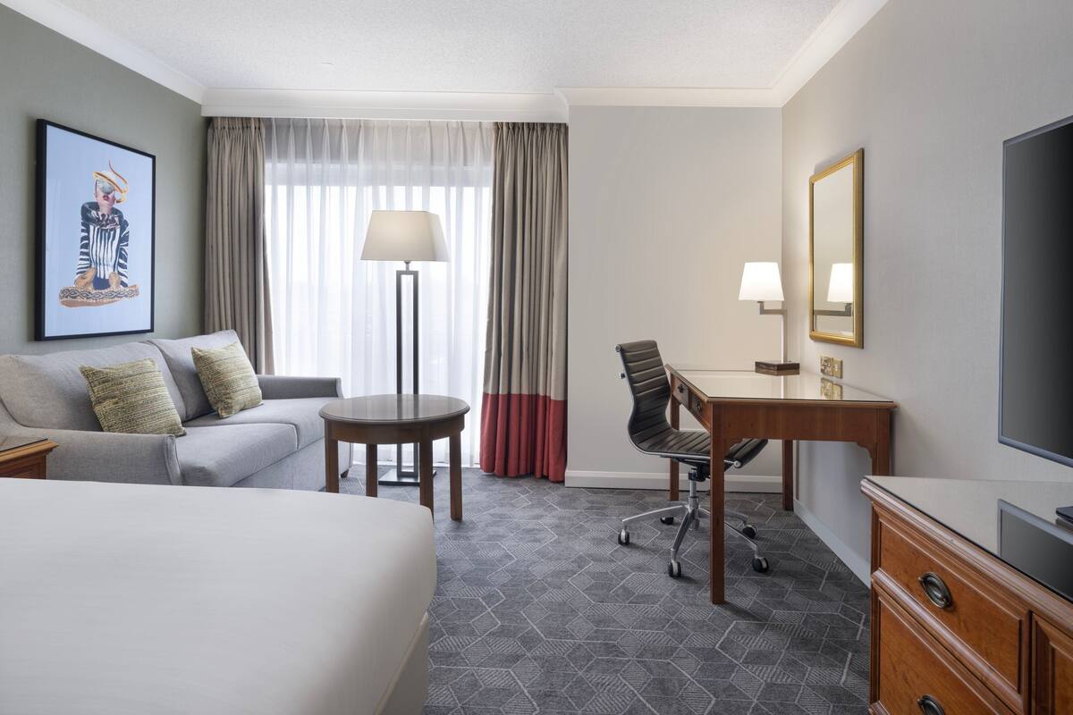 A hotel room with a bed, a desk, a chair, a sofa, a TV, and a painting on the wall.