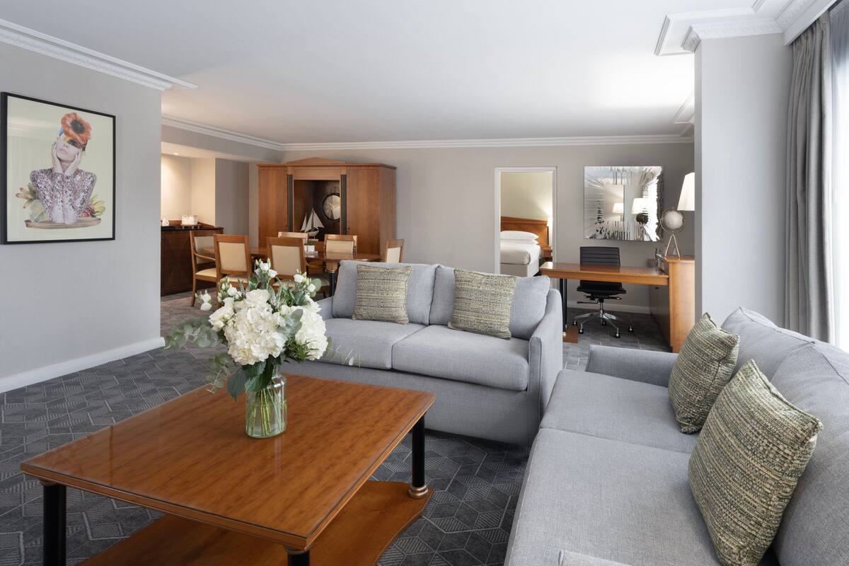 A modern hotel room with a grey sofa, a wooden coffee table, and a dining area with a wooden cabinet and chairs.