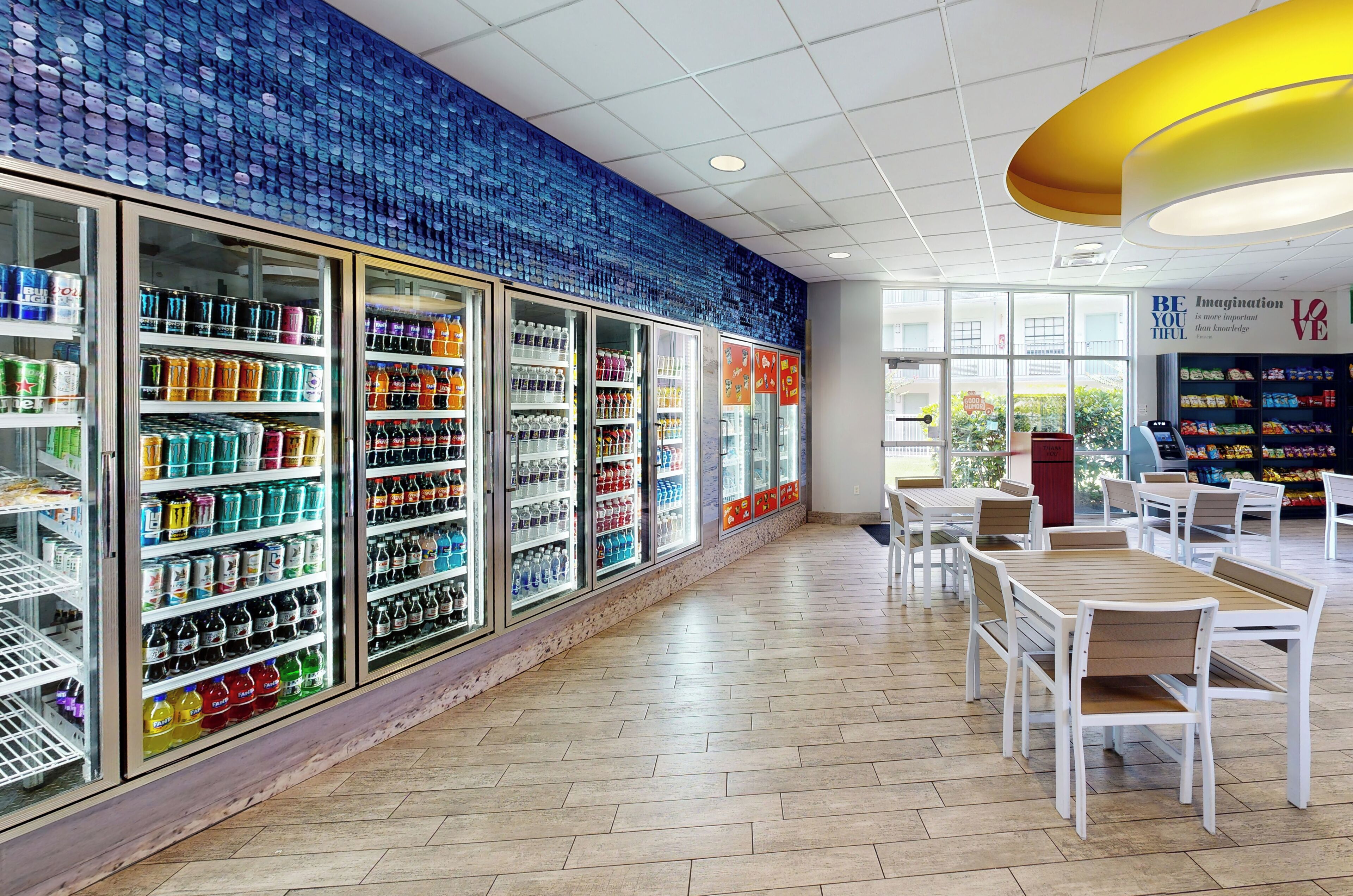 Ein modernes Interieur mit gekühlten Getränkespendern, weißen Tischen und Stühlen sowie einer blauen Mosaikwand.