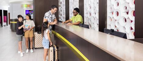 A hotel reception area with a modern design, including a reception desk, a concierge desk, and a wall with circular patterns. There are several guests standing and interacting with the staff.