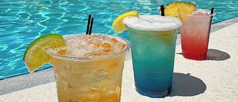 A poolside with three colorful drinks, a clear blue pool, and a building with balconies and umbrellas in the background.