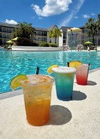 Una piscina con tre bevande colorate, acqua cristallina e un edificio sullo sfondo con balconi e ombrelloni.