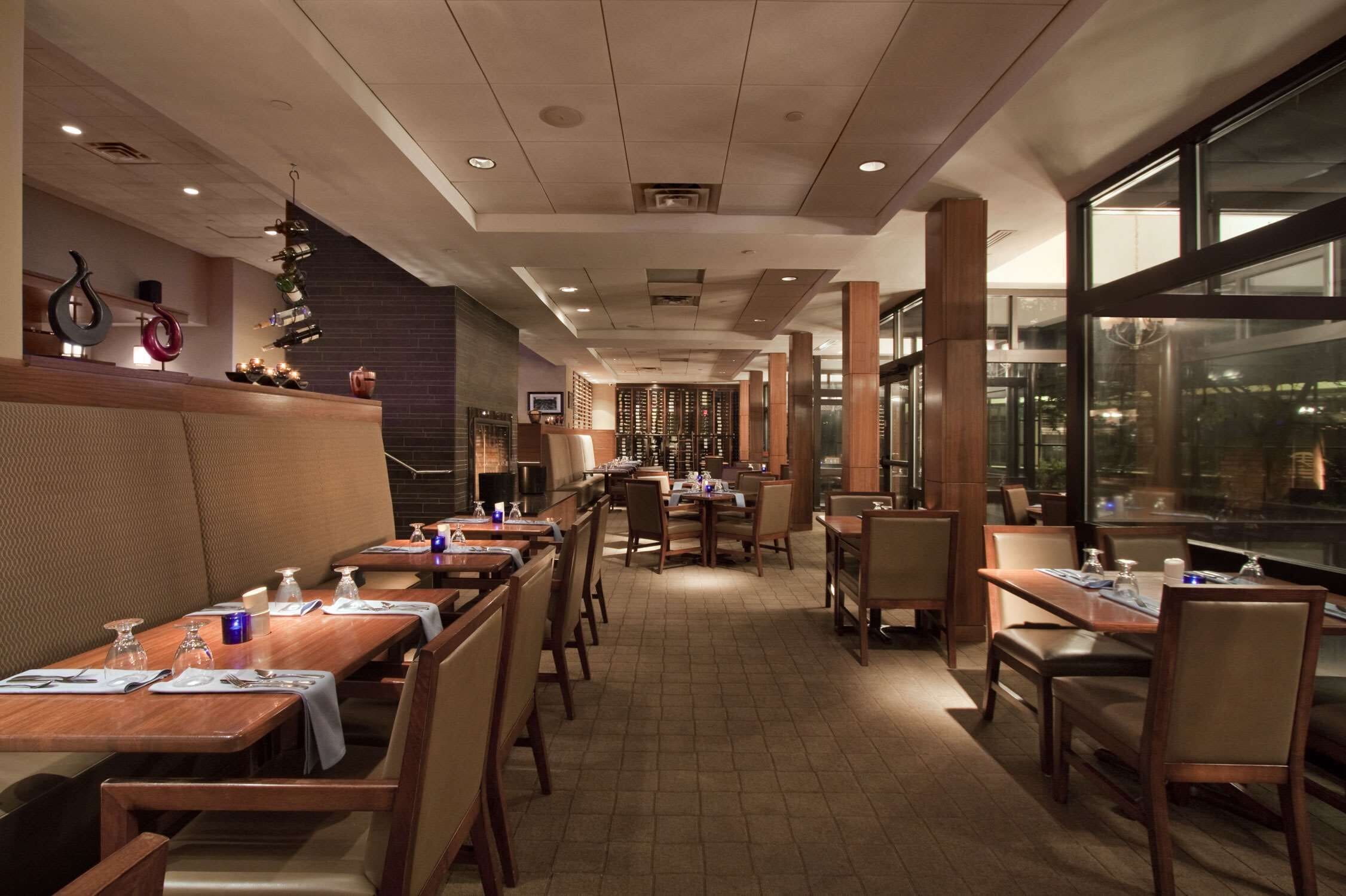 A restaurant with long wooden tables and booths, a brick wall with a wine rack, and large windows offering a view.