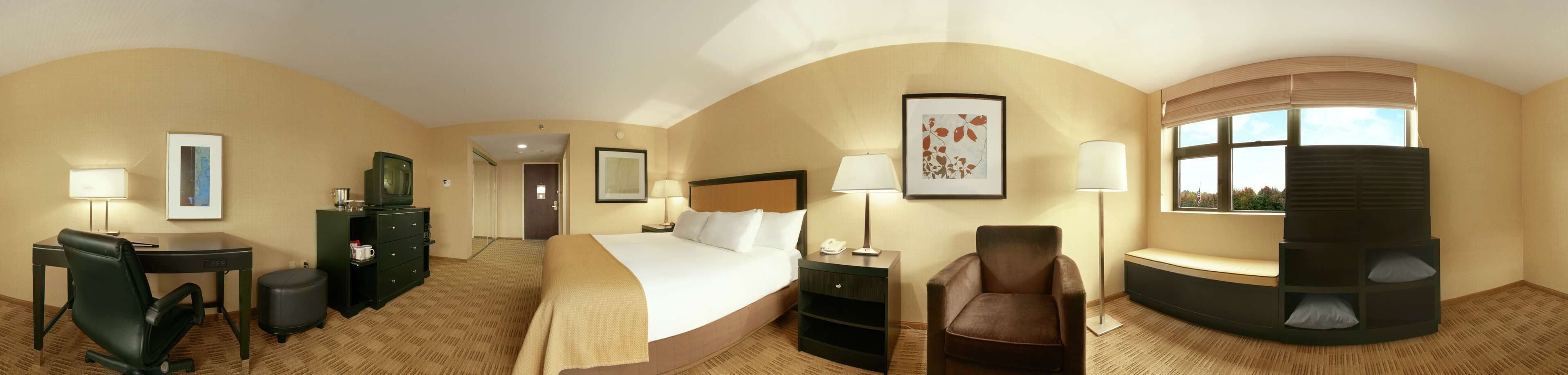 A hotel room with a bed, desk, chair, bedside table, lamp, and a view of the outdoors.