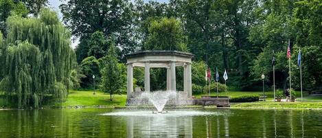 A gazebo with a fountain in the center, surrounded by a pond and trees.