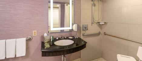 A bathroom with a sink, mirror, towel rack, shower, and a wheelchair-accessible chair.