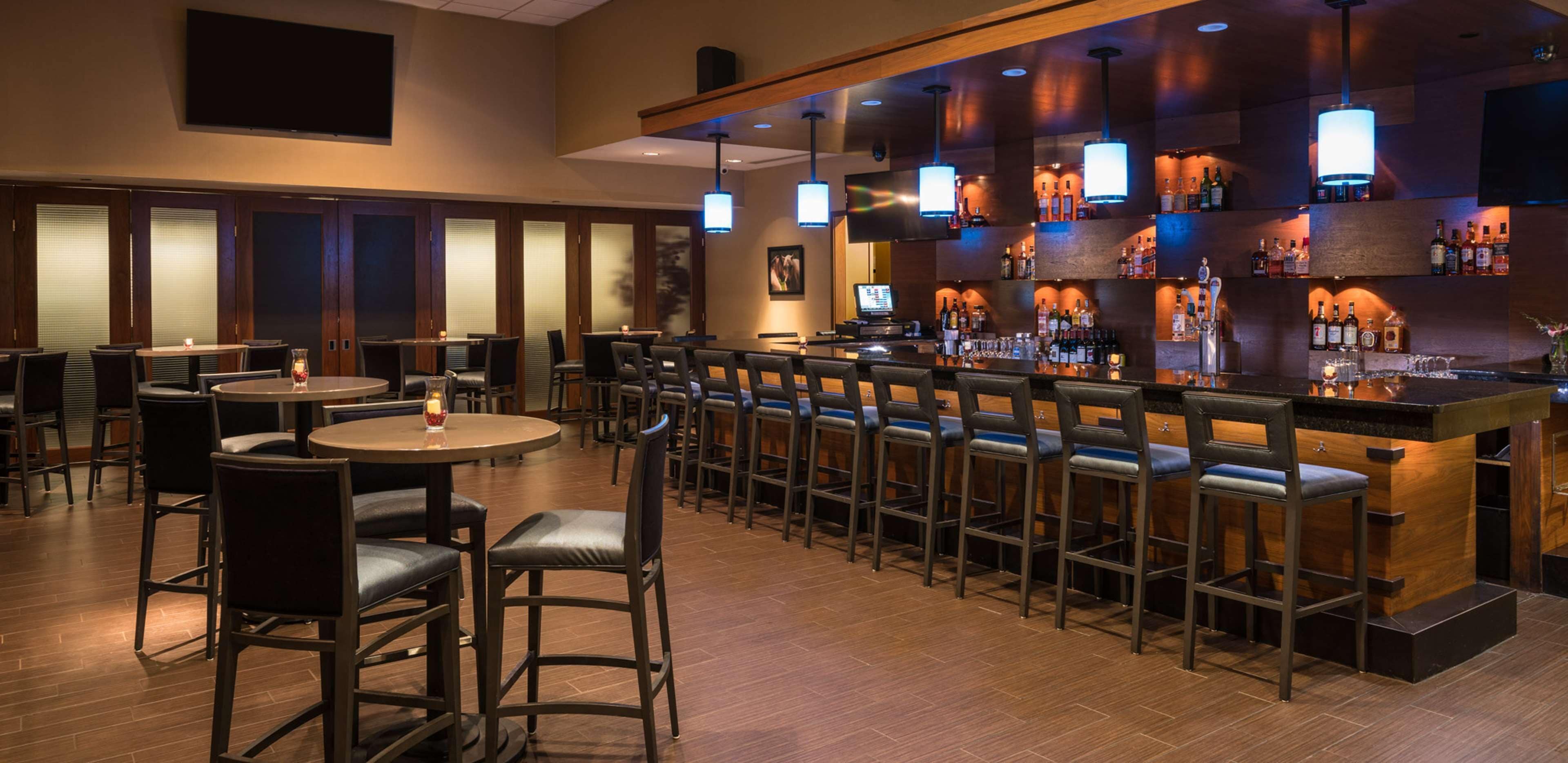 Un espace bar offrant différentes options de sièges, notamment des tables rondes et des tabourets de bar. On y trouve plusieurs luminaires suspendus et un comptoir de bar où sont exposées diverses bouteilles et verres.