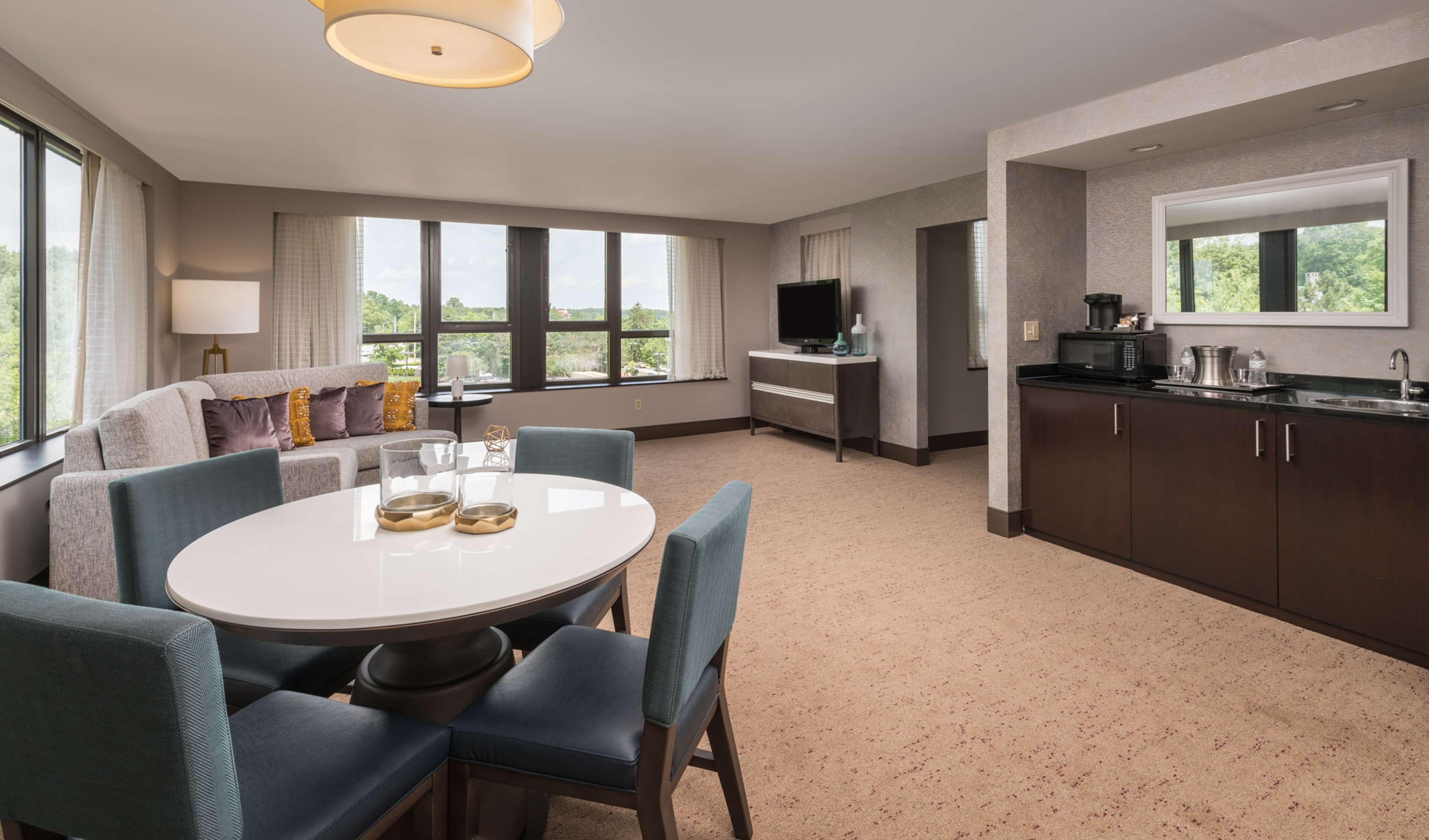Et moderne hotelværelse med et rundt spisebord, blå stole, en sofa med puder, et fjernsyn, et tekøkken med mikroovn og store vinduer med udsigt til grønne omgivelser.