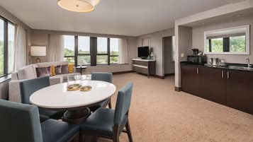 Una habitación de hotel moderna con una mesa redonda, sillas azules, un sofá con cojines, un televisor, una zona de cocina con microondas y ventanas grandes que dan a zonas verdes.