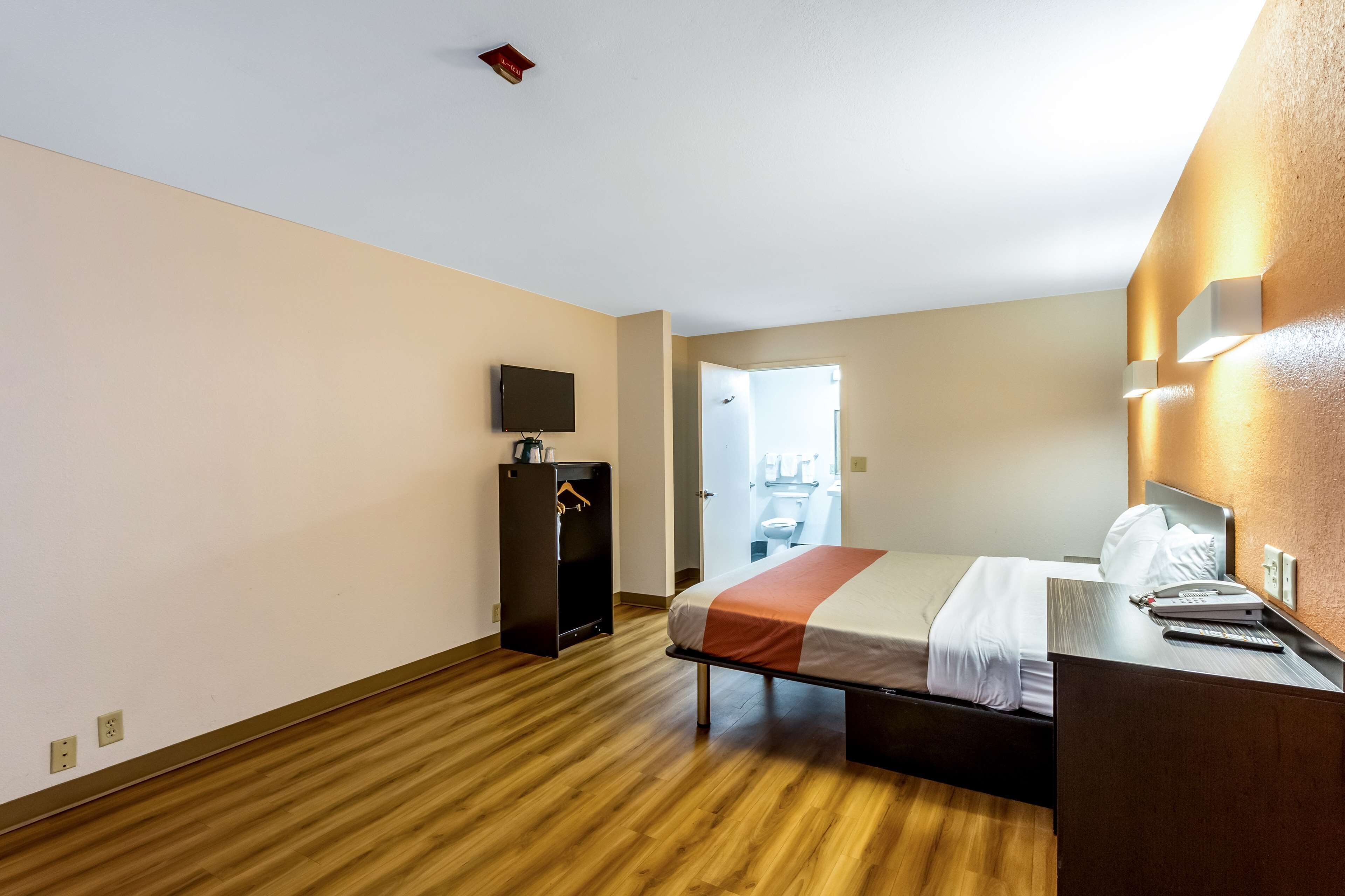 Une chambre d’hôtel avec un plancher en bois, un lit recouvert d’une courtepointe blanche et orange, un bureau avec un ordinateur, un téléviseur fixé au mur et une armoire.