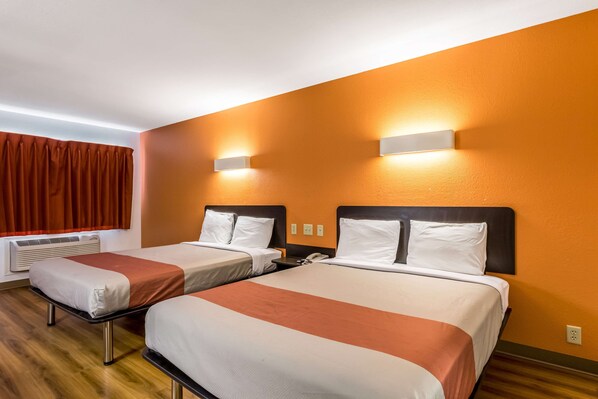 A hotel room with two beds, each with white linens and a red accent, a wooden floor, and an orange wall.