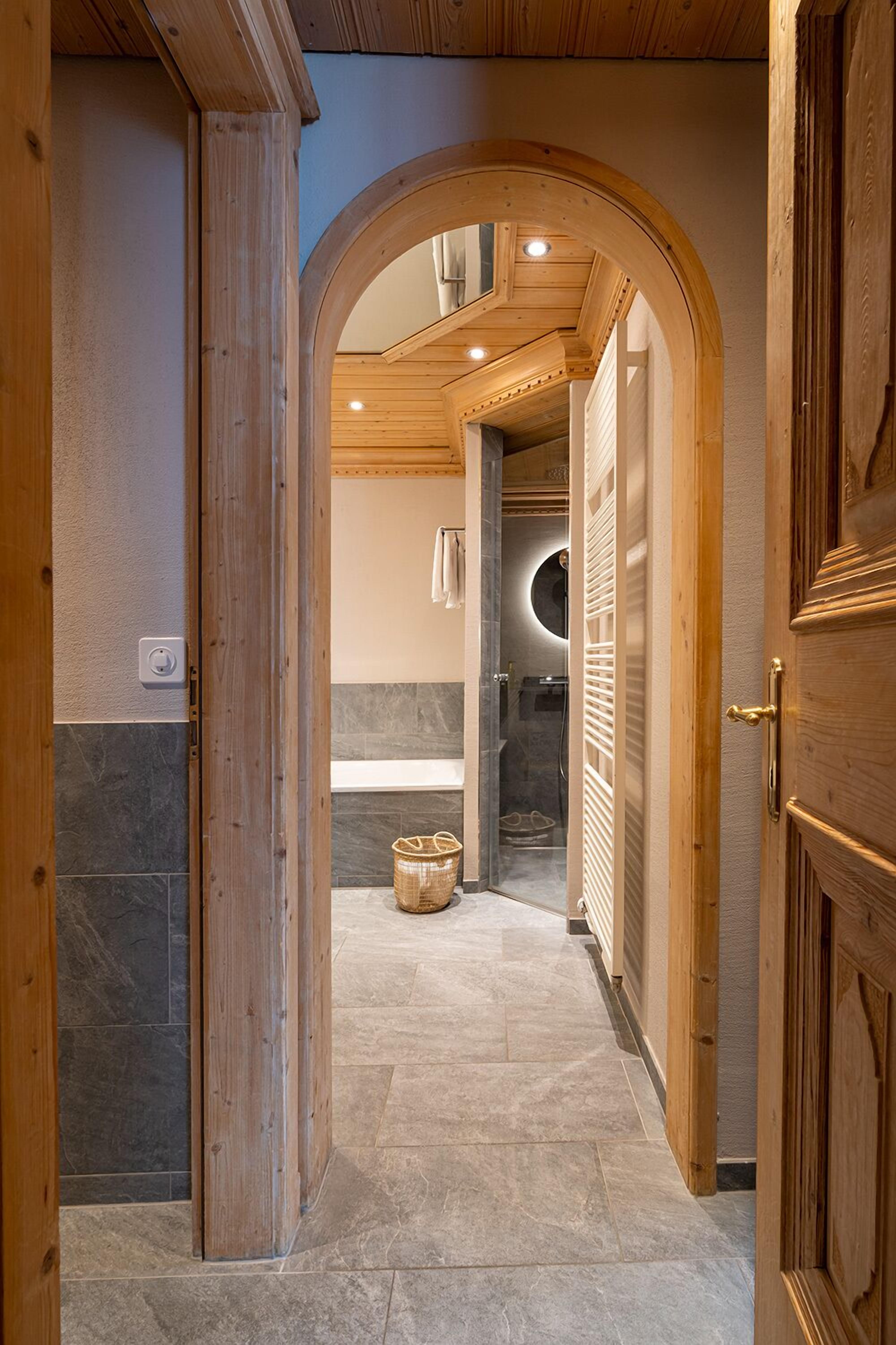 A hallway with wooden doors, a stone-tiled floor, and a view into a bathroom with a circular mirror and a towel rack.