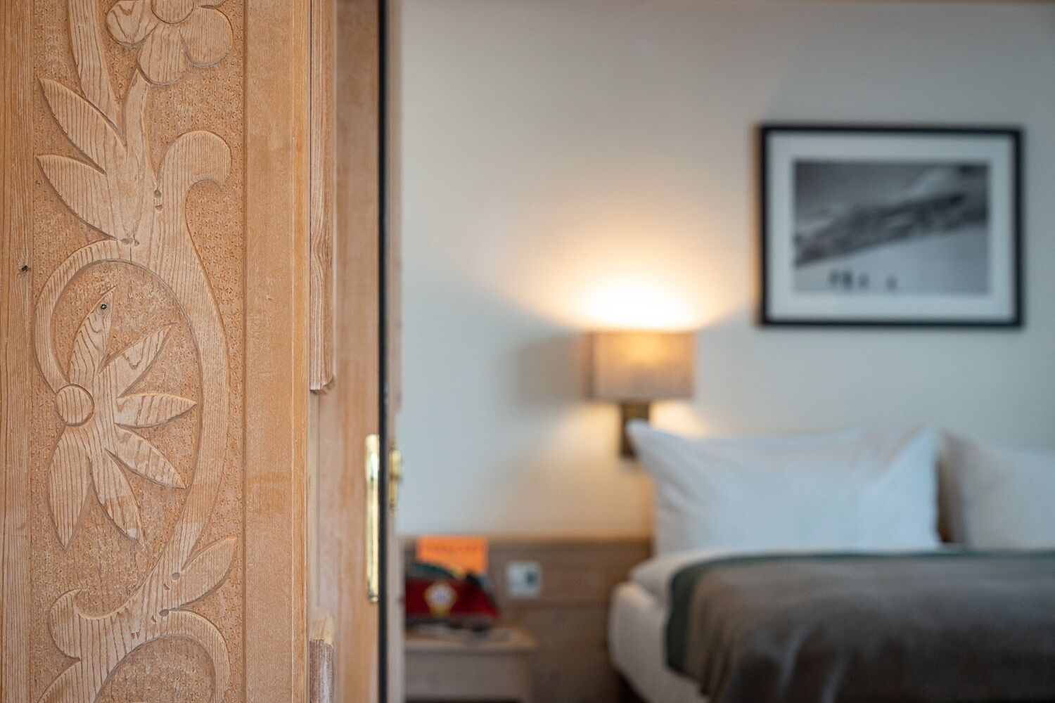 Ein Hotelzimmer mit einer Holztür, verziert mit floralen Schnitzereien, einem Bett mit weißer Bettwäsche, einem Nachttisch mit Lampe und einem gerahmten Bild an der Wand.
