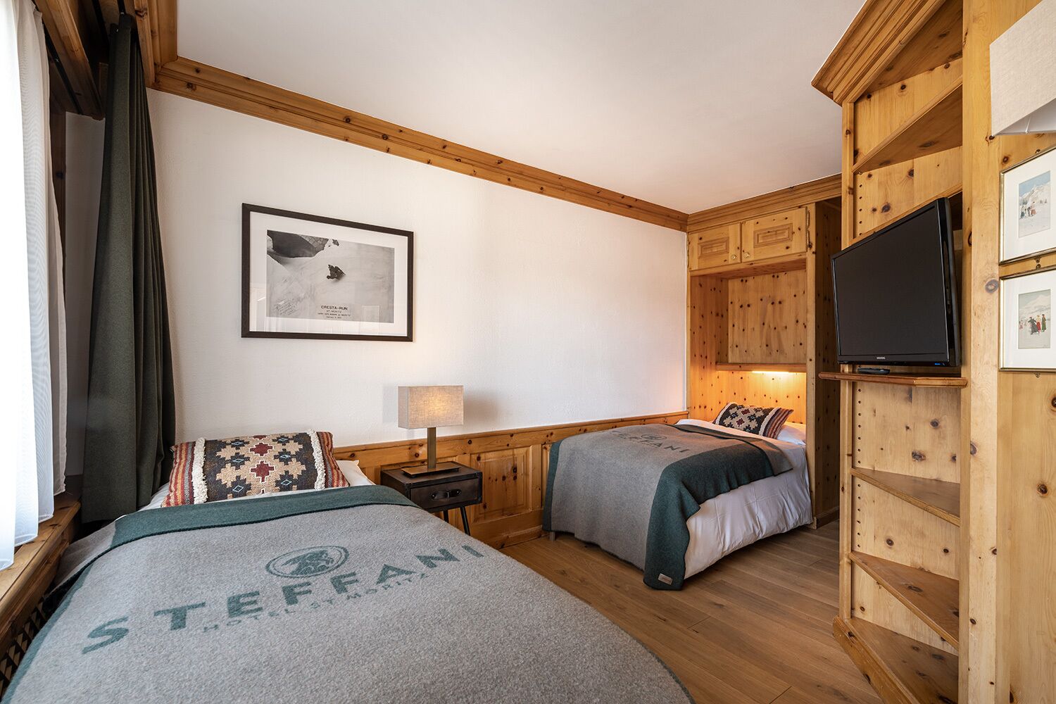 A hotel room with two beds, wooden paneling, a mounted TV, a framed picture on the wall, and a lamp on a bedside table.