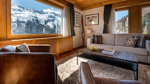 A cozy living room with a wooden ceiling, large windows with mountain views, a sofa, a coffee table, and a fireplace. - Hotel Steffani (St. Moritz)