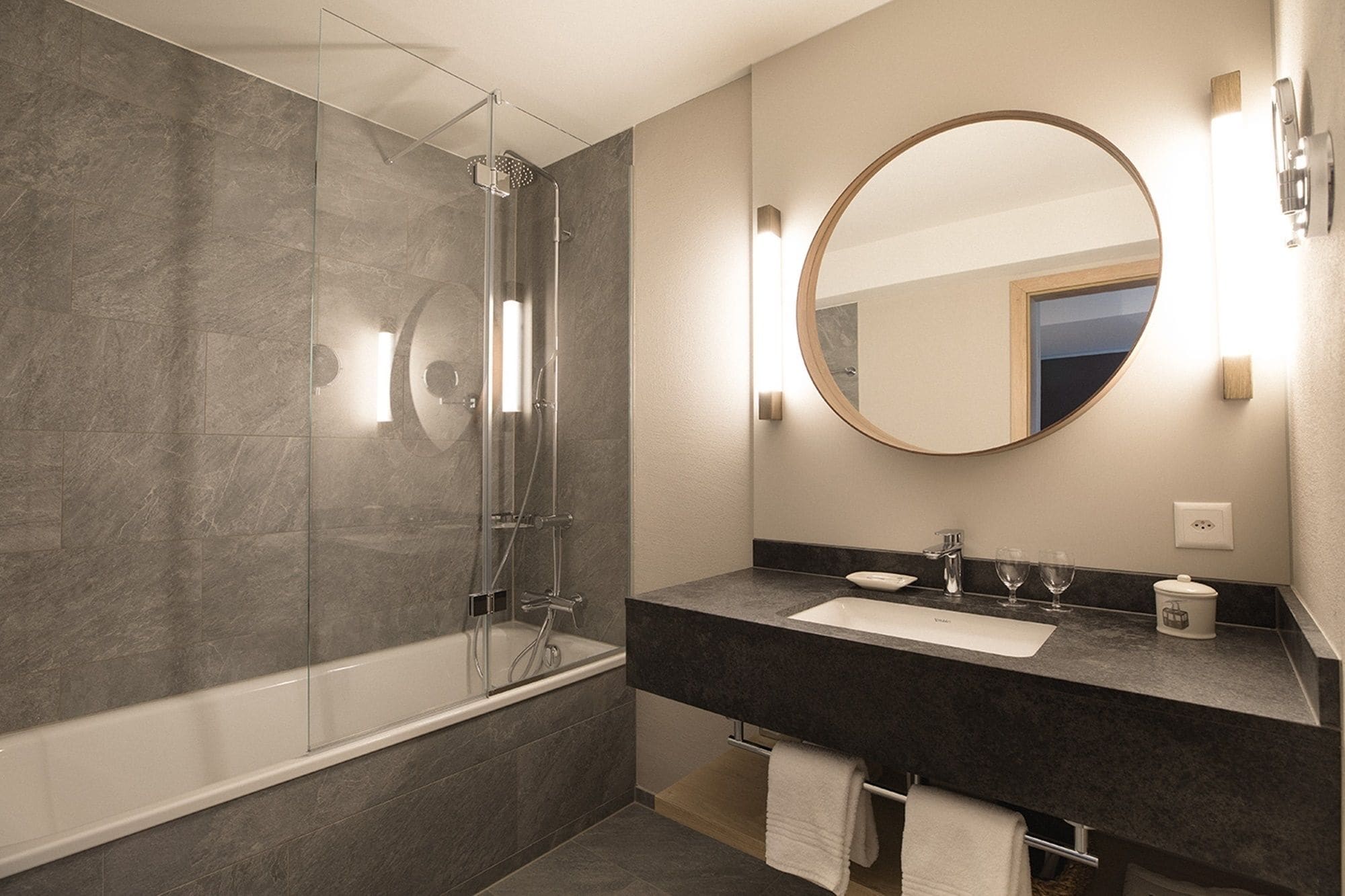 Un baño moderno con ducha envuelta en vidrio, una bañera, un lavabo con tocador y un espejo redondo.