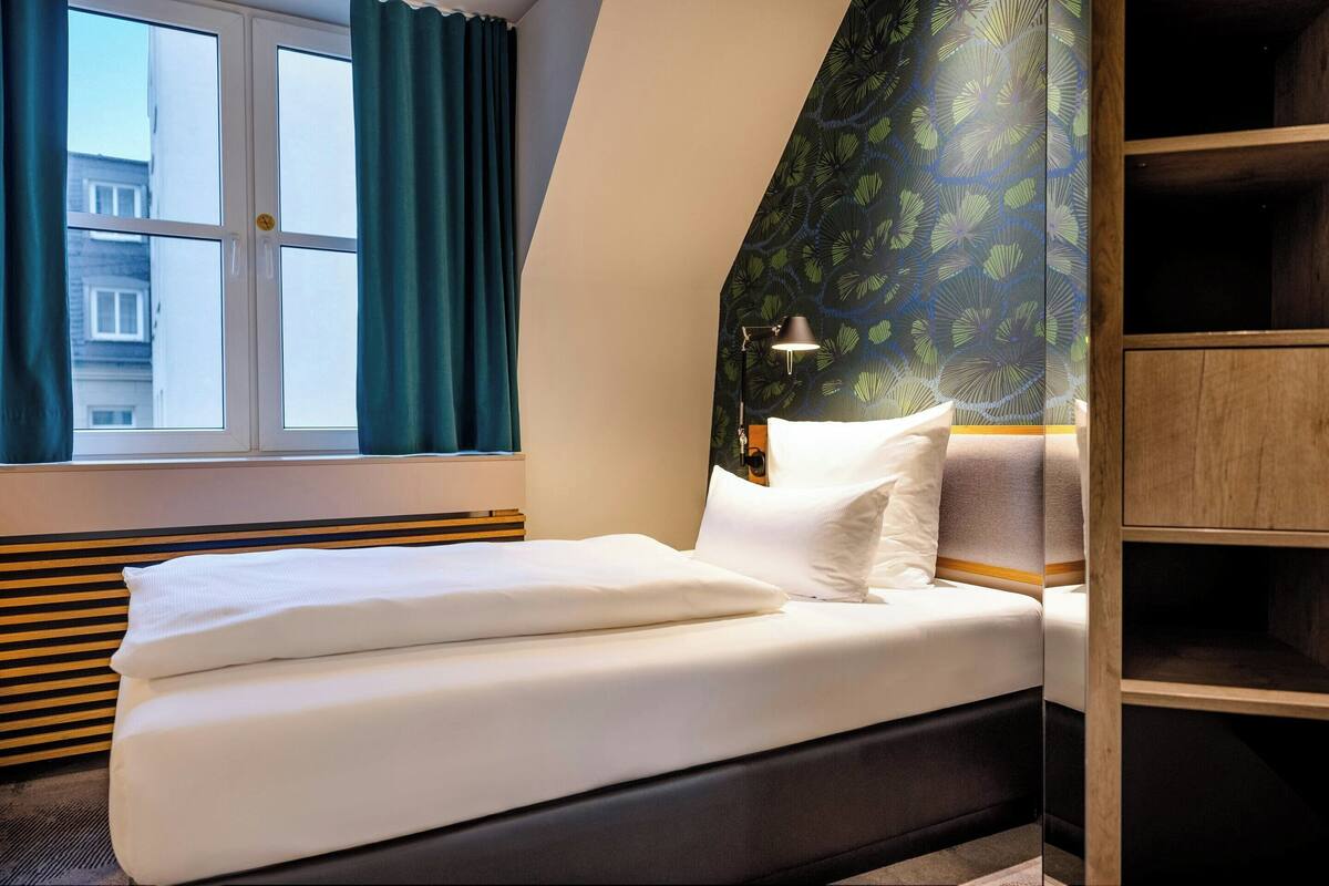 A neatly made bed with white linens, a window with teal curtains, and a patterned wall.