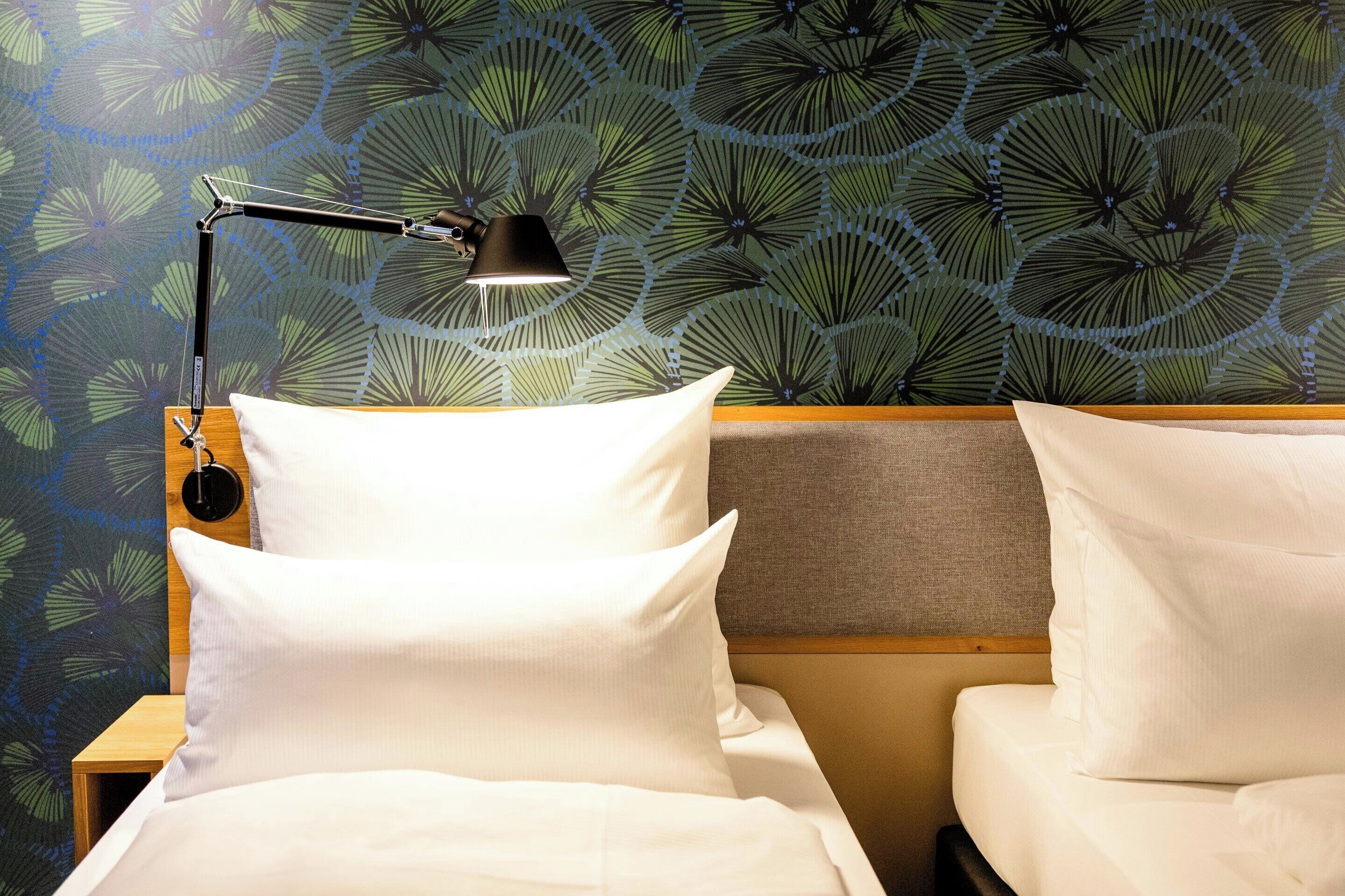 Quarto de hotel com duas camas, uma mesa de cabeceira, uma luminária e papel de parede estampado.