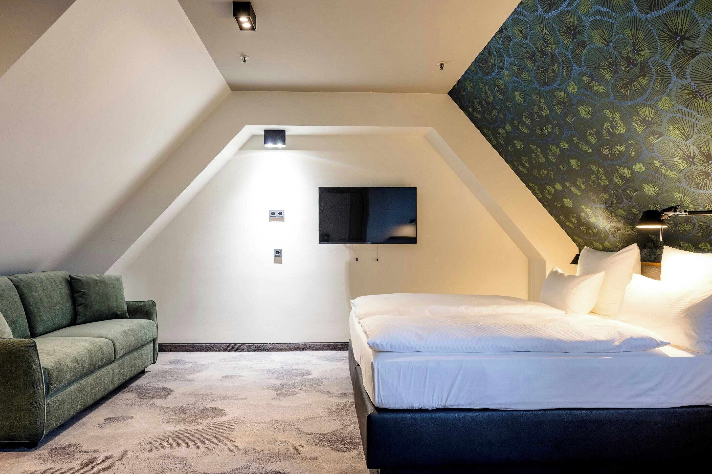 Un dormitorio con cama, sofá verde, televisor de pantalla plana montado en la pared y techo estampado.