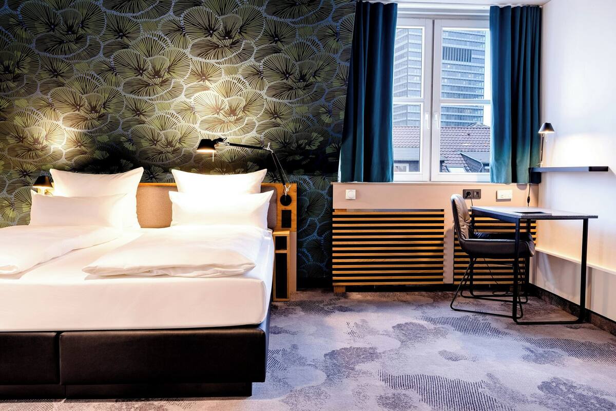 Una camera d'albergo con un letto, una scrivania, una sedia, una finestra con tende e carta da parati decorata.