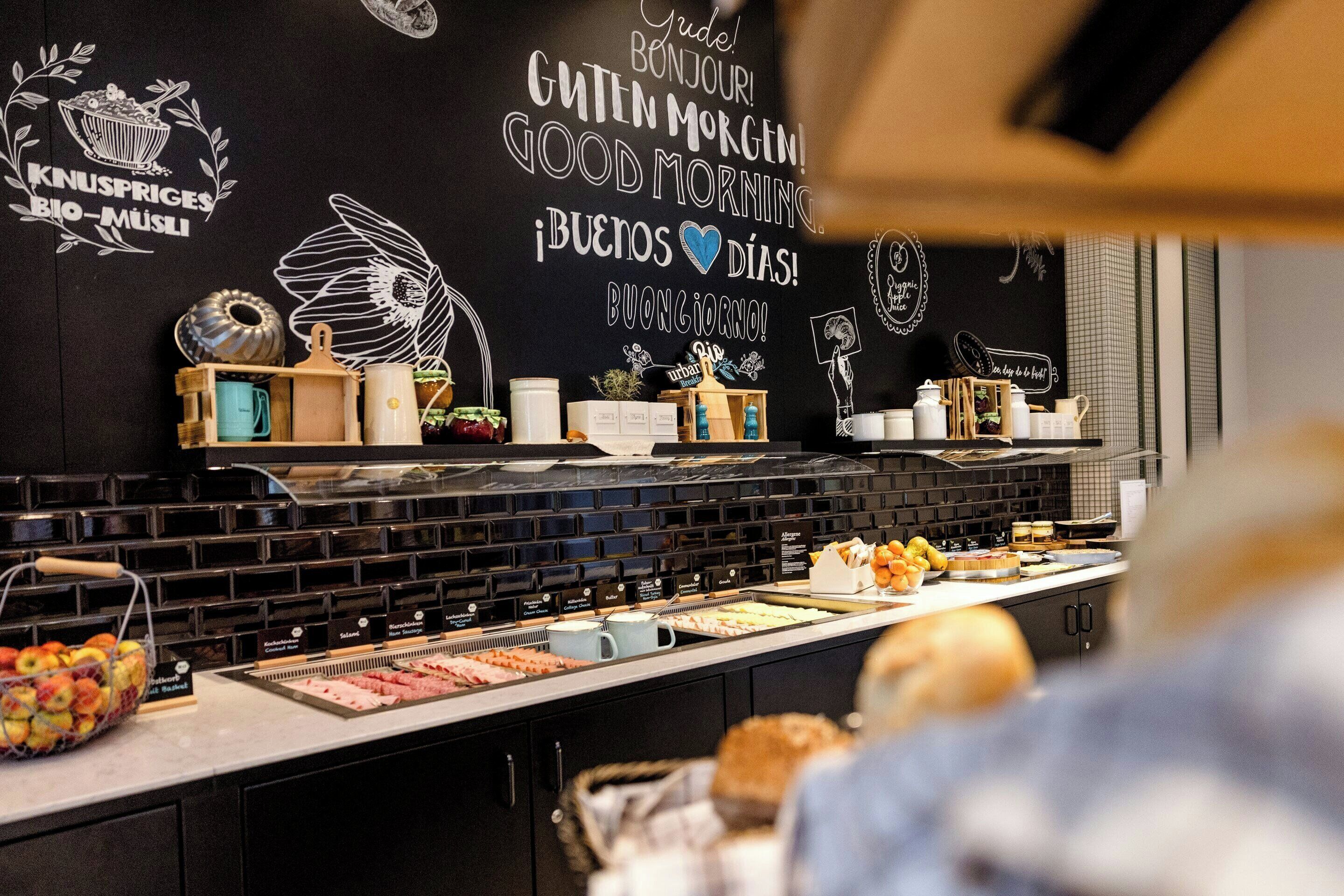 A breakfast buffet with various food items and a black wall with multilingual greetings.
