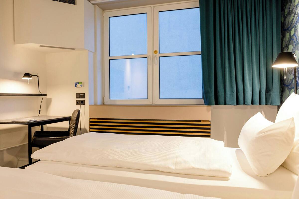 Camera d'albergo con un letto, una scrivania, una sedia, una finestra con tende e una lampada.