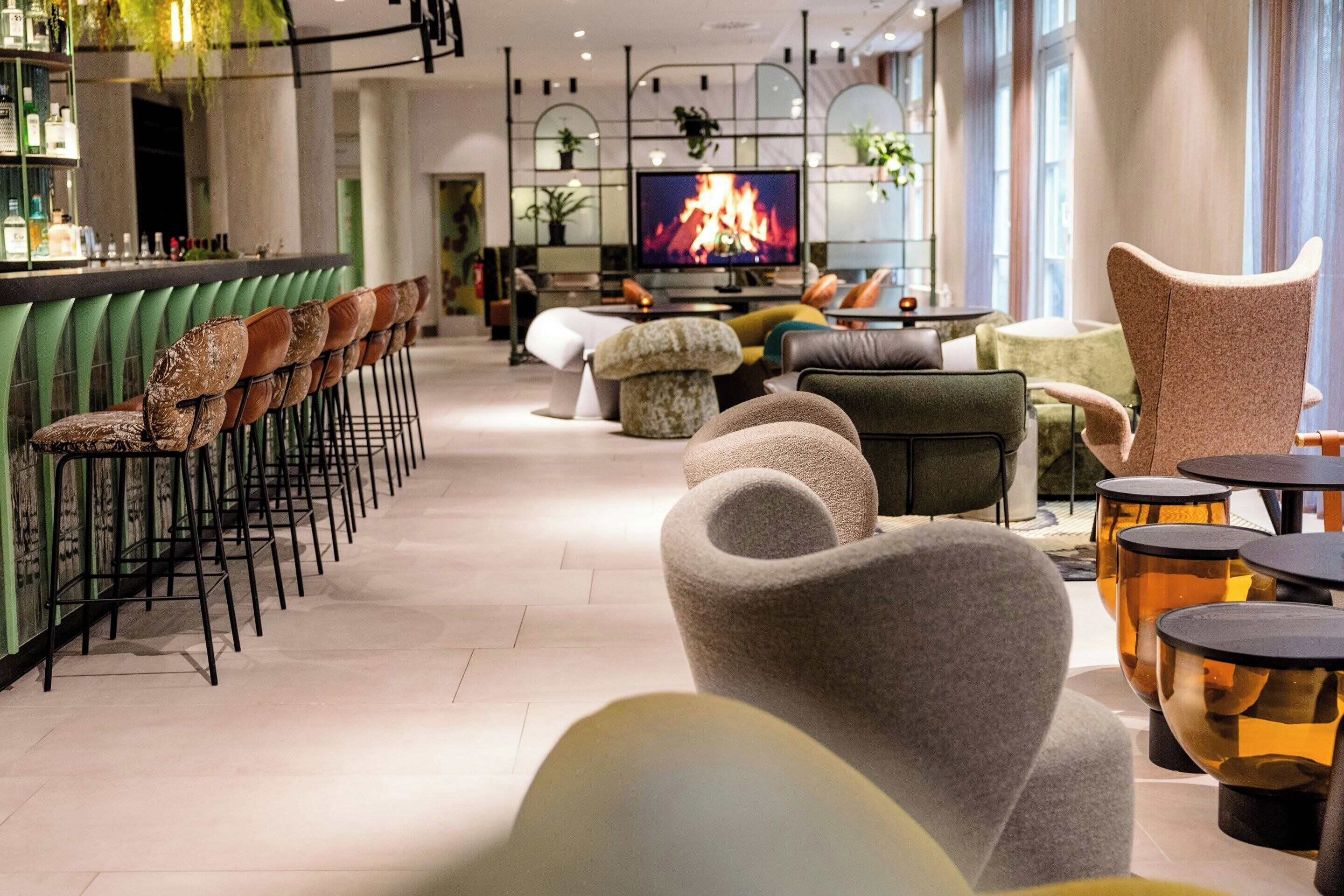 Eine moderne Bar mit grünen Barhocker, einem Fernseher, der ein Feuer zeigt, und einer Lounge mit Sofas und Poufs.