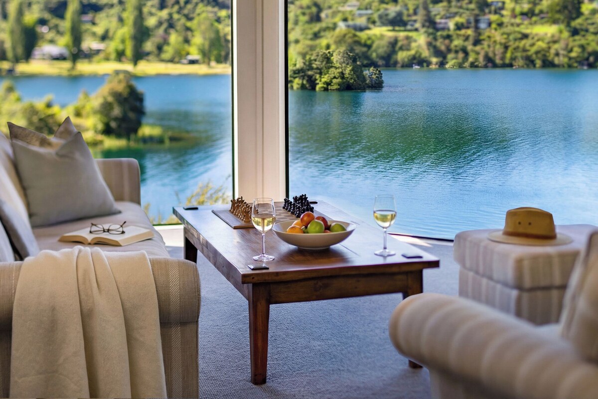 A room with a view of a lake, a wooden table with a bowl of fruit and wine glasses, and a sofa with a book and glasses on it.