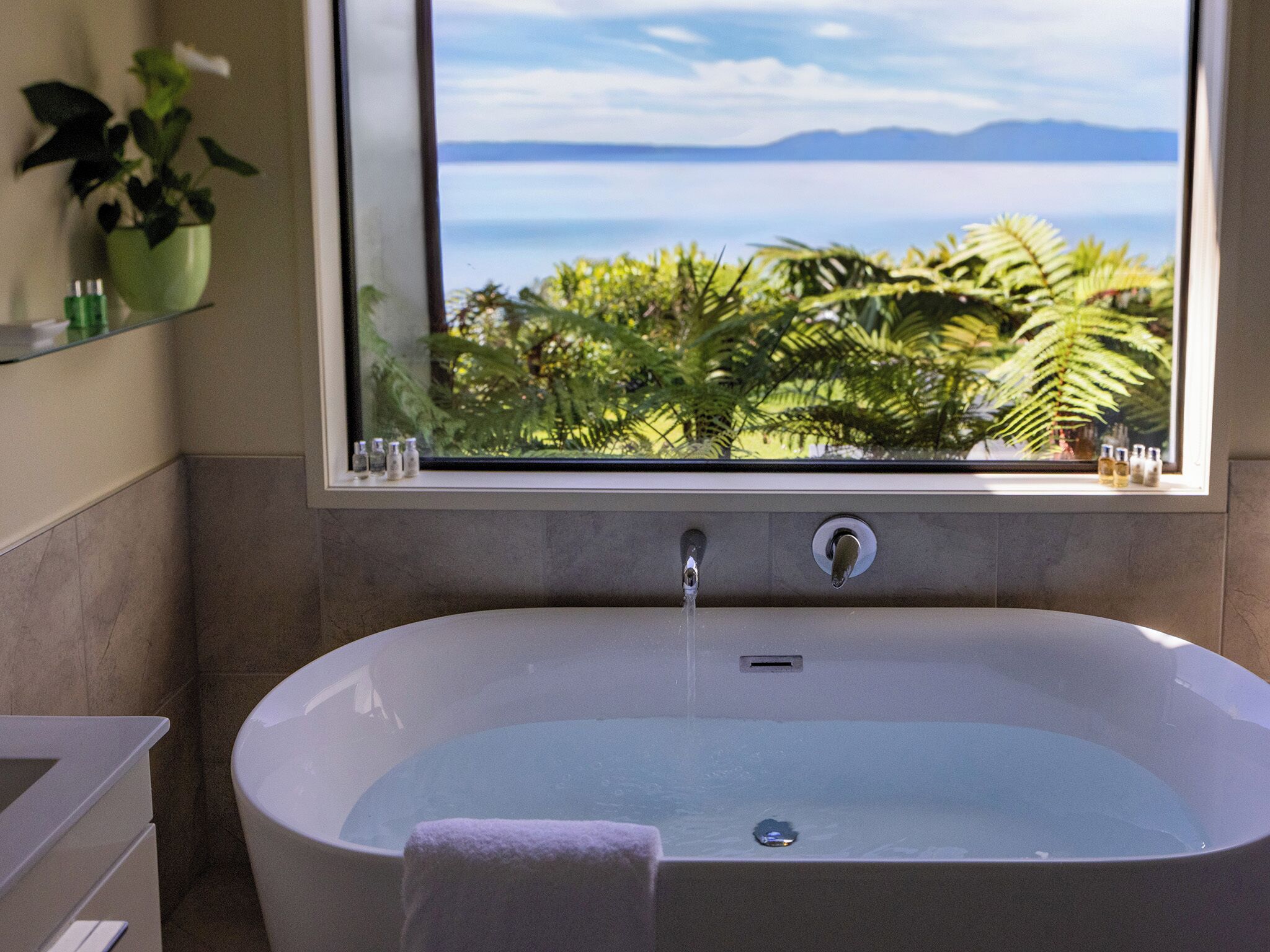 Un baño moderno con una bañera independiente, una ventana con vistas a un paisaje tropical y al mar, y un estante con artículos de aseo.