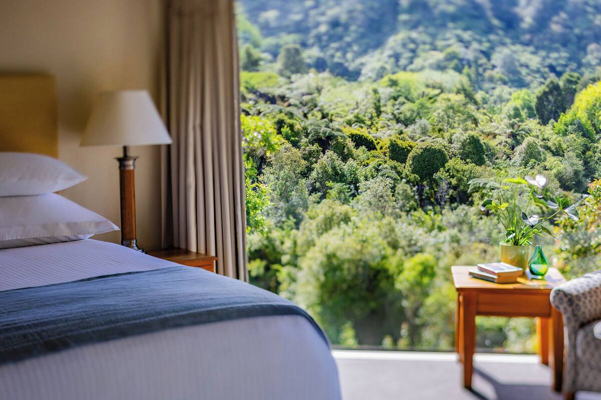 A hotel room with a bed, a nightstand, a lamp, a bedside table with a book and a vase, and a view of a lush green landscape.