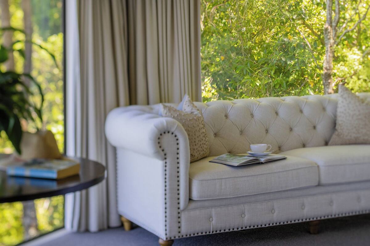 A white tufted sofa with a cushion, a small table with a book and a cup, and a view of greenery through a window with curtains.