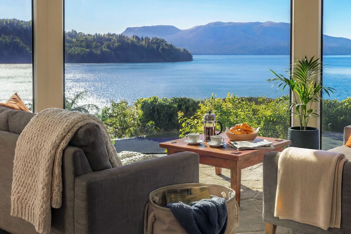 A cozy living room with a large window overlooking a lake and mountains. There is a sofa, a coffee table with a basket of bread, and a potted plant.
